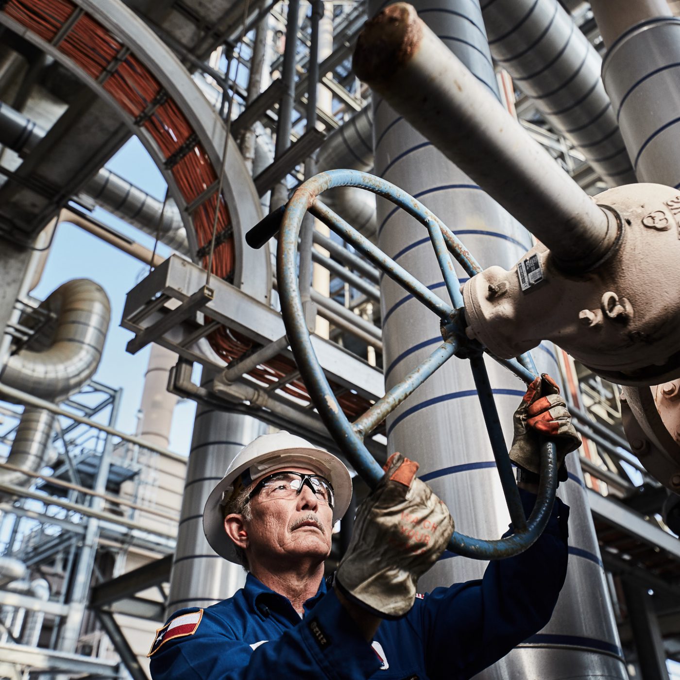 A man wearing personal protective equipment is working on a valve in an industrial oil refinery.